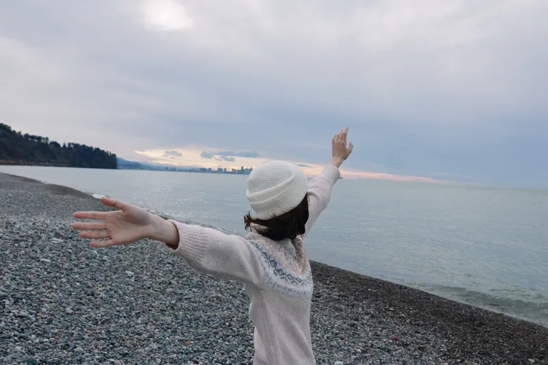 Beach scene with a person on a pebble shore, arms outstretched to the sea and horizon, wearing a light sweater and hat, conveying freedom and peaceful coastal mood.