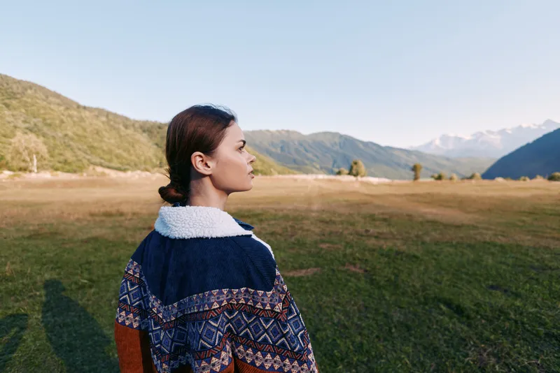 Woman in patterned sweater stands in meadow looking at distant mountains, profile view in outdoor landscape. Nature travel scene with calm light, open field and valley retreat.