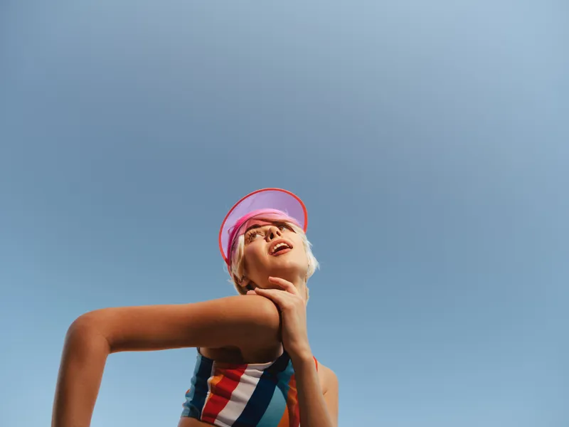 A child wearing a striped swimsuit and pink visor gazes upward against a clear blue sky, capturing a moment of carefree summer wonder and athletic confidence.