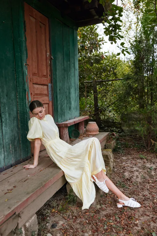 A woman in a pale yellow dress sits on a wooden porch in a rustic outdoor setting, leaning slightly, enjoying a calm sunny moment with soft breeze and vintage charm.
