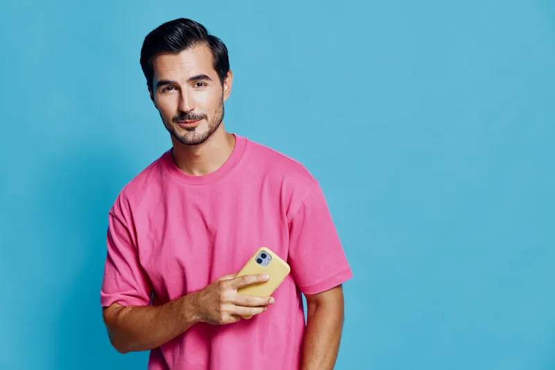 Full body smiling cheerful young man wearing pink t-shirt holds mobile phone in hand, looking confidently at camera on solid blue background. People lifestyle concept