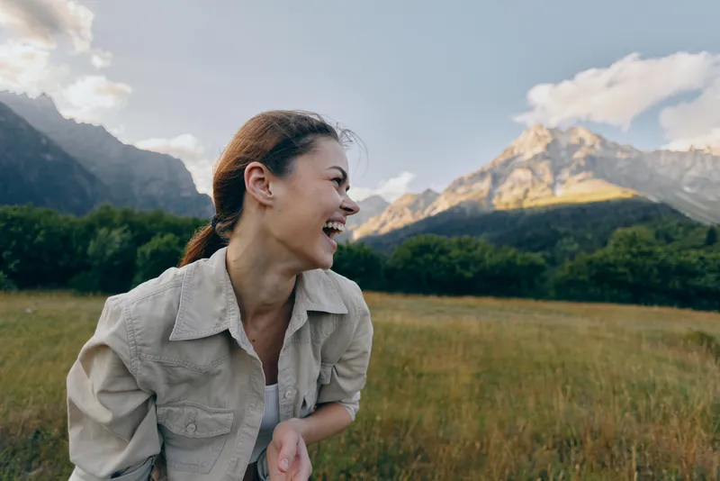 A laughing woman stands in a sunlit grassy field with a dramatic mountain landscape behind her, enjoying a carefree outdoor moment and a sense of freedom.