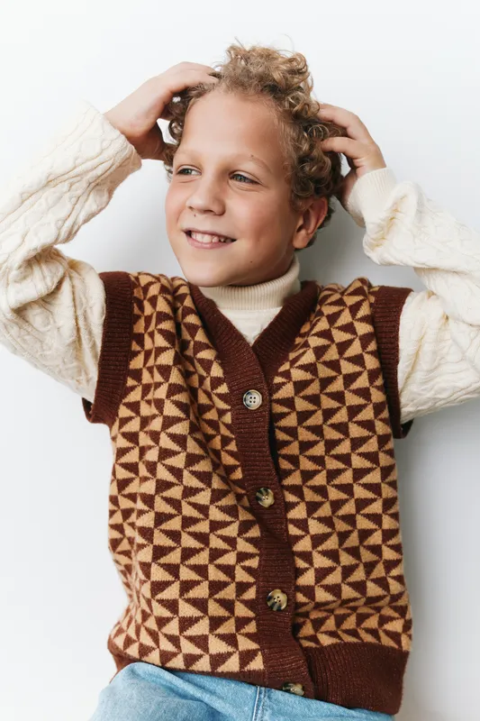 Joyful young boy poses against a plain backdrop, wearing a geometric brown and beige knitted vest over a cream turtleneck, conveying warmth, innocence, and a candid playfulness.