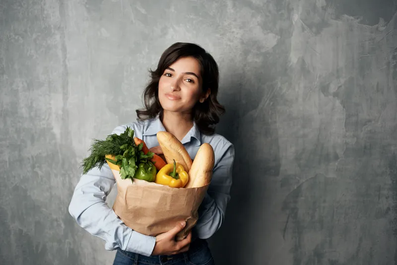 Улыбающаяся брюнетка с продуктовой сумкой из супермаркета, полной овощей, на тему диетического питания