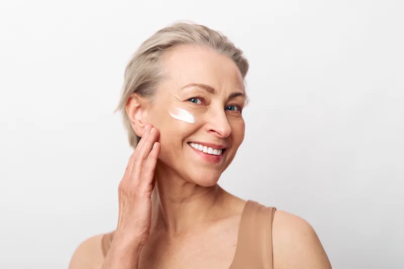 Smiling mature woman applying face cream on cheek shows skincare routine and healthy skin care concept with natural beauty on white background in bright soft lighting.