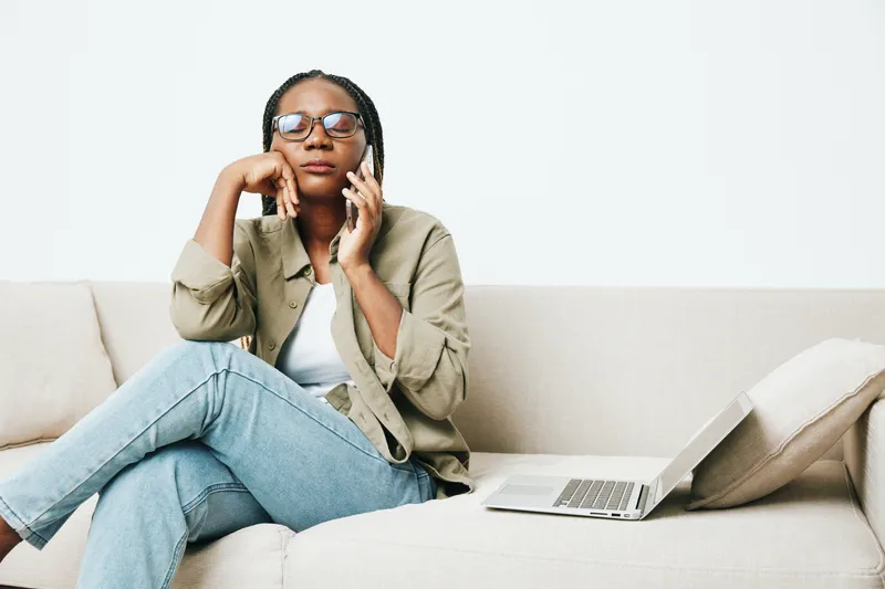 Стильная афроамериканская женщина-фрилансер работает, сидя на диване дома с ноутбуком и телефоном