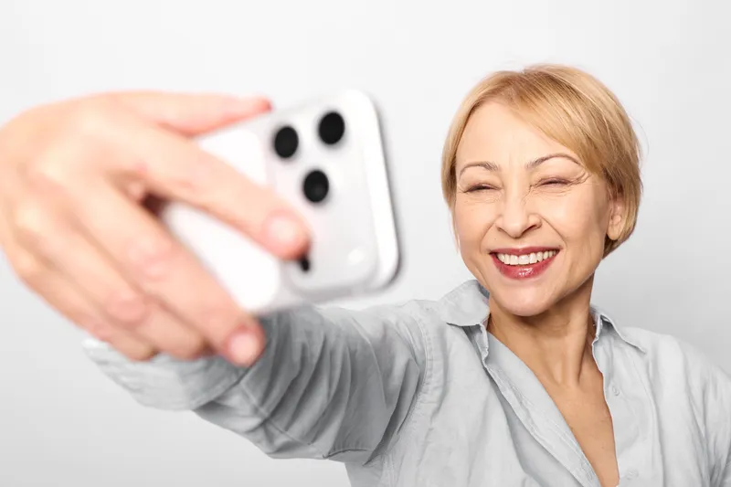 Senior woman selfie smartphone smiling hand technology communication indoors, she raises a phone for a cheerful portrait, soft light and casual outfit suggest wellbeing and connection