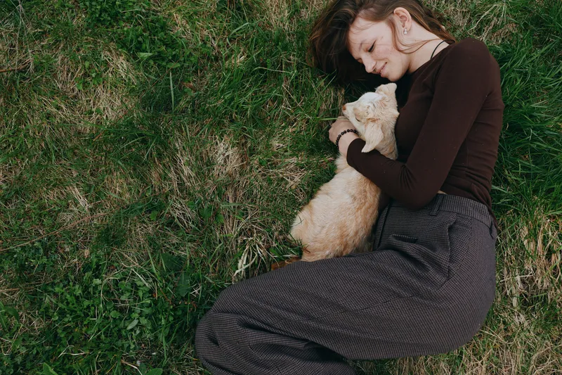 Lifestyle moment of a woman resting on grass, embracing a calm goat kid, showing tenderness, comfort, connection, and warm sunlight in an outdoor natural setting, gently, quietly, and happily.