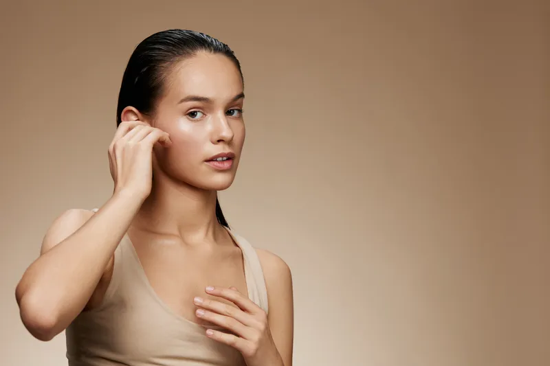Portrait of a young female model with wet hair, wearing a beige top, showing a natural look against a soft brown background, conveying serenity and confidence
