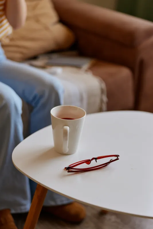 Relaxed woman in a cozy living room sits on a sofa with a white mug on a round table and a pair of glasses nearby, showcasing a casual home lifestyle and tranquil mood.