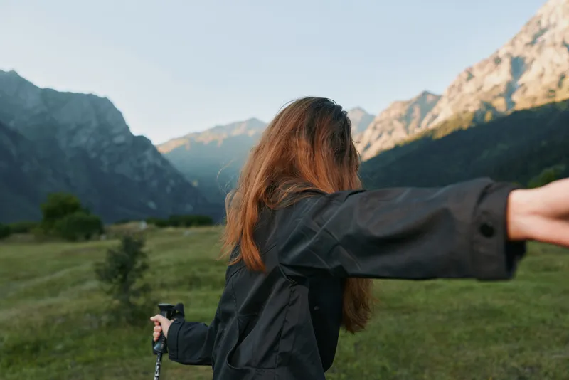 A lone hiker stands in a wide meadow with rolling hills and distant peaks, arms outstretched in freedom, wearing a dark jacket and holding a trekking pole.