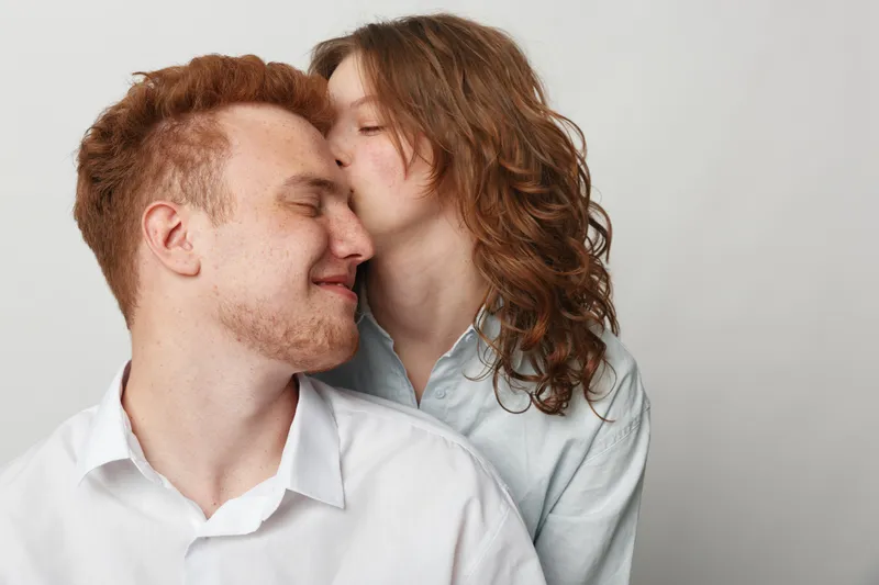 Young couple in casual shirts share a tender moment as woman kisses man on cheek. Intimate love and affection concept with soft natural light and neutral background.
