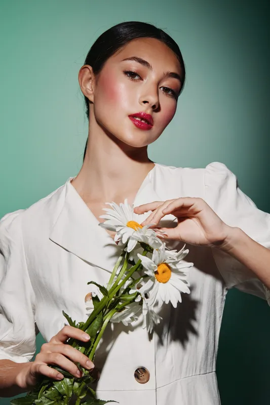 Portrait of a stylish young woman with glossy red lips and glowing skin holding white daisies. Soft lighting enhances natural beauty and elegance in a fashion inspired setup.