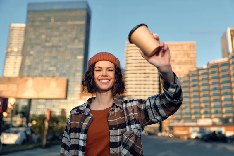 Woman holding coffee cup, smile in urban morning scene, candid lifestyle portrait capturing authenticity and golden hour glow. Emotional storytelling of mindful living and joy.