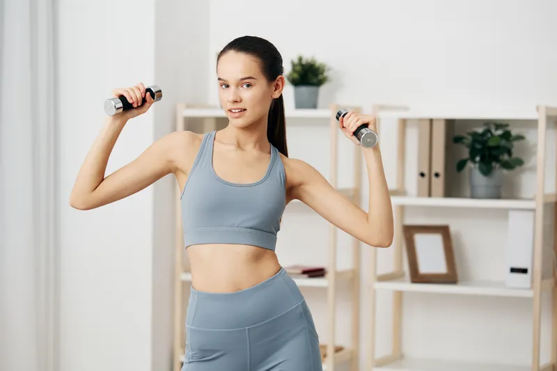 A woman in light blue athletic wear performs a dumbbell shoulder press at home, showcasing woman fitness, sport, workout and training with focused energy in a bright room.