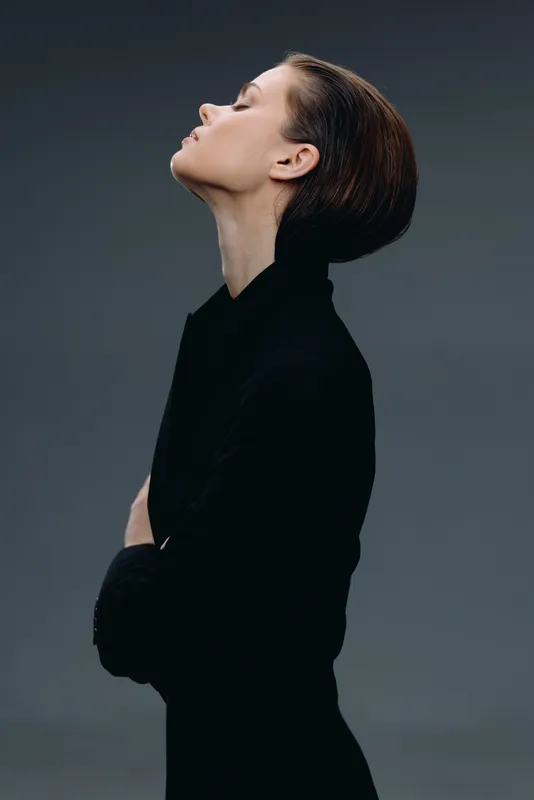 Elegant woman in black turtleneck with closed eyes posing on dark background, showing profile and calm expression.