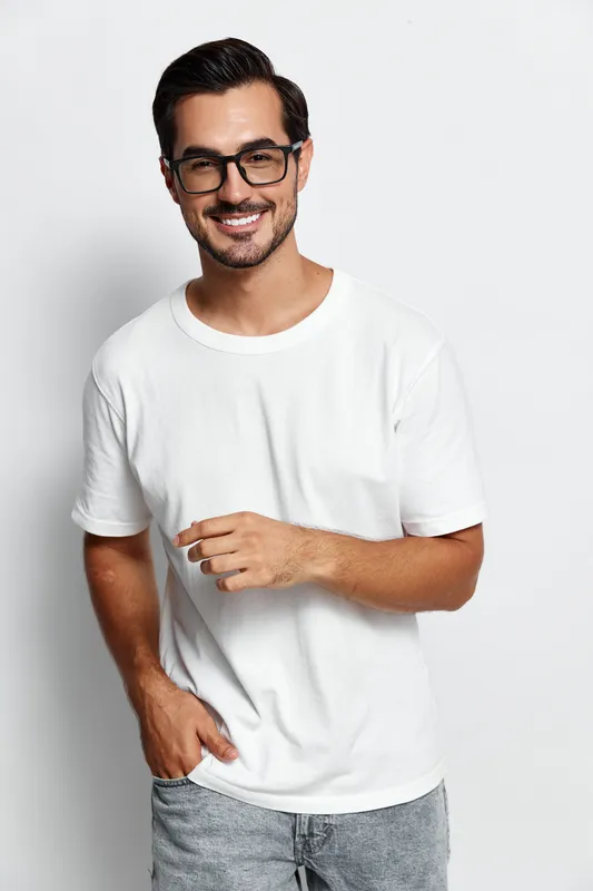 Smiling young man with glasses, wearing a white t-shirt, confidently poses for the camera while standing against a plain background. People lifestyle concept.
