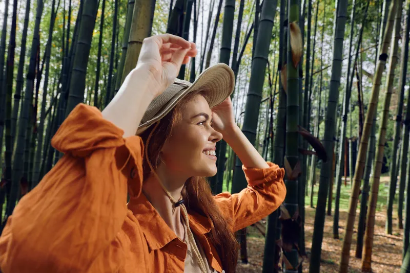 Woman bamboo forest hat smile nature portrait in sunlight, young traveler in orange shirt enjoying a peaceful walk and leisure exploration among tall green trees and fresh foliage.