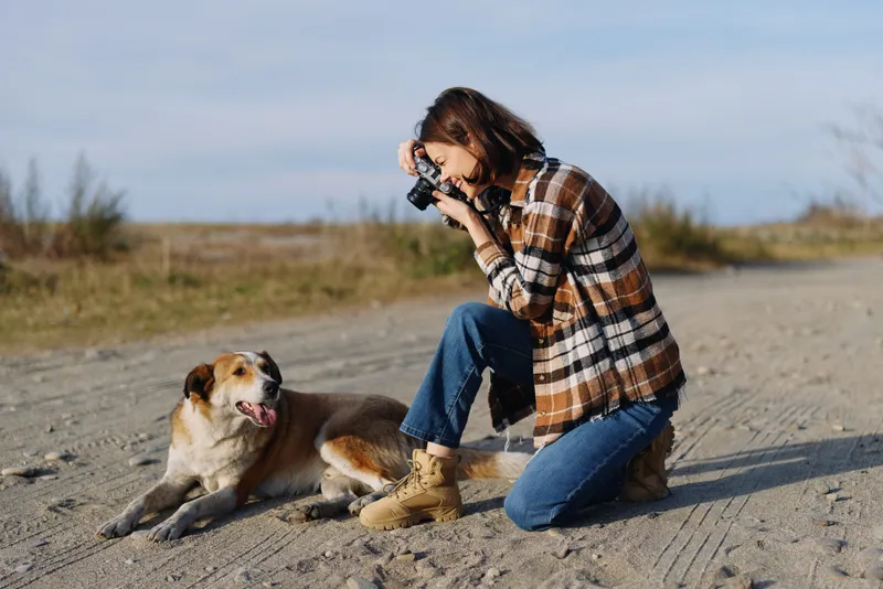 Photographer kneels on a sandy path with a camera, capturing a friendly dog in an outdoor landscape under clear sky and open space for a natural moment and candid lifestyle shot.