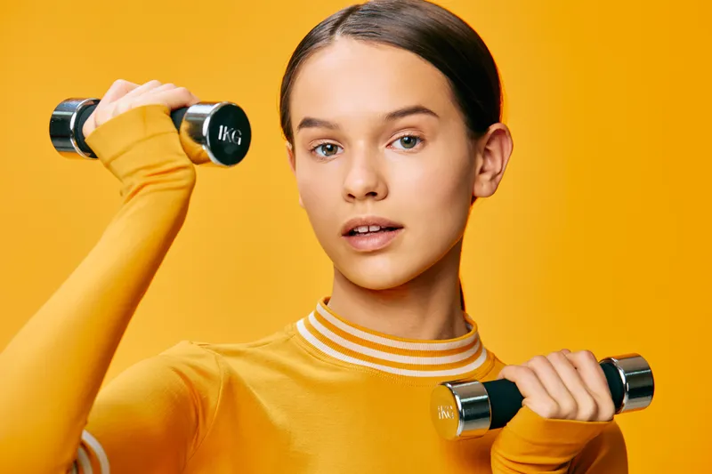 Woman fitness sport training with dumbbells in hand, exercising on bright orange studio backdrop, healthy lifestyle and strength building, focused expression and athletic style.