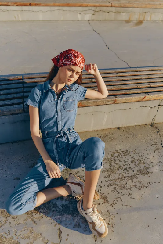 Lifestyle woman in modern boho-western denim street style wearing casual sneakers and a red bandana, captured with warm film color tones and natural sunlight outdoors.