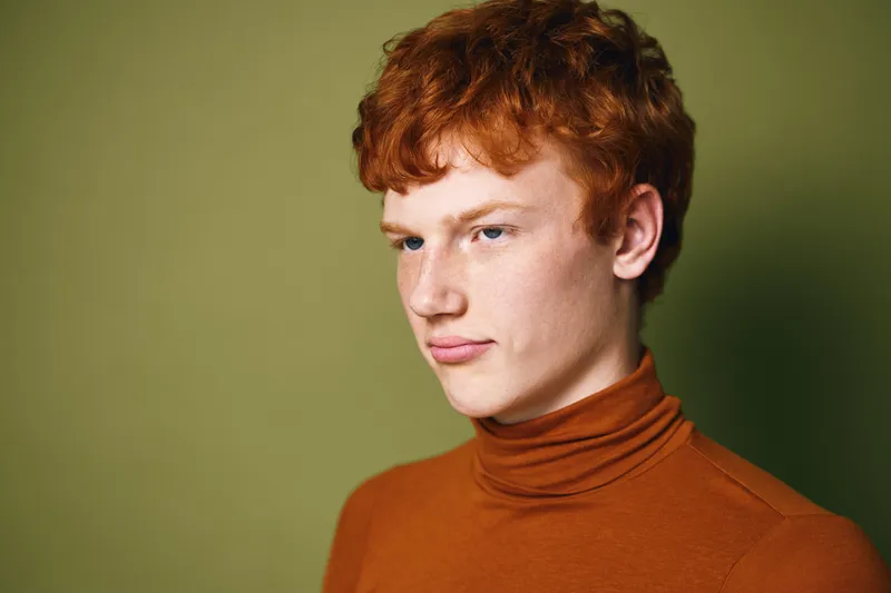 Portrait of young man with red hair wearing orange turtleneck looking away with thoughtful expression against olive green background. Closeup of male face and curly hairstyle indoors.
