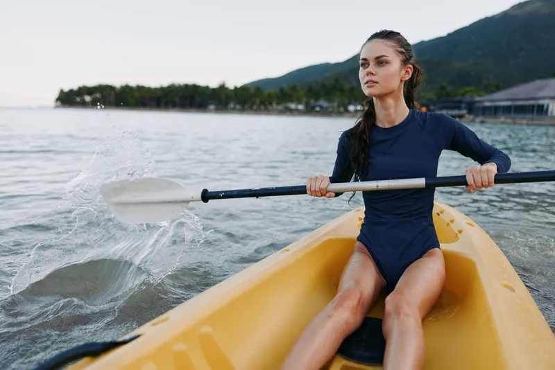 A focused female kayaker in a dark blue suit paddles a yellow kayak across tranquil water, creating splashes as the oar slices the surface.