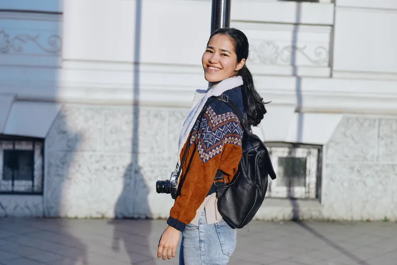 Woman smiling outdoors with camera, backpack, and sweater, walking on a sunny street, showcasing casual urban style and everyday photography energy
