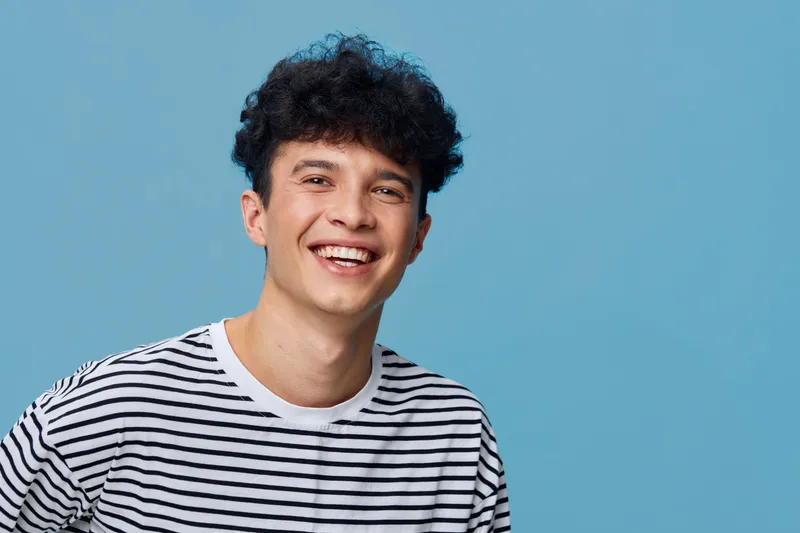 Smiling young man wearing a striped shirt against a blue background, expressing happiness and friendliness in a casual portrait with bright lighting.