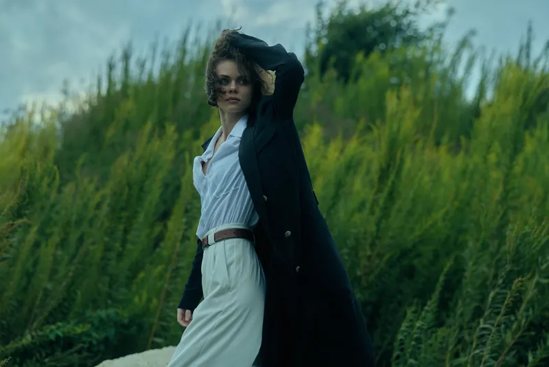 Elegant woman in a flowing white dress beneath a long dark coat, posing in a windy field with hand in hair, stylish and dramatic mood in nature.