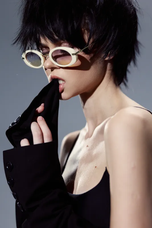 Stylish woman wearing white sunglasses and black clothing biting sleeve with moody expression in studio lighting.