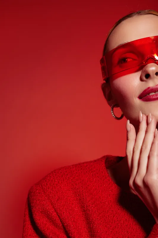Close-up of a stylish woman wearing red sunglasses and a red sweater, posing with hands near face against a red background. Concept of fashion, style, and confidence.