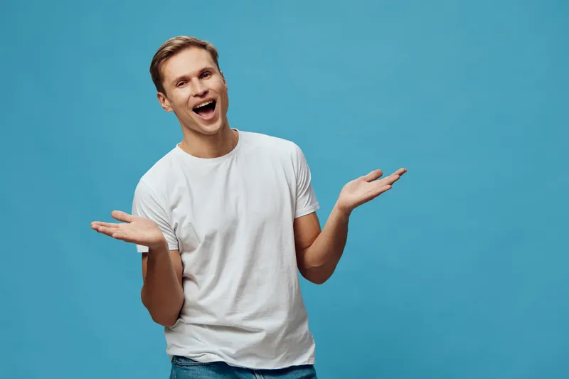 Smiling young man with light brown hair wearing a plain white t-shirt looking cheerful and playful, gesturing with open hands, isolated on a bright blue background, casual lifestyle shot, positive moo