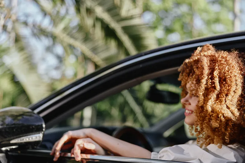 smiling woman with curly hair sitting in car, enjoying sunny day outdoors with green palm tree background, relaxed and happy mood