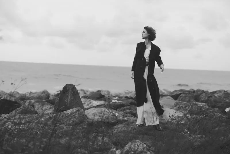 Woman standing on a rocky shoreline in a long dark coat, wind in her hair, sea and clouds behind, moody black and white fashion scene, solitary figure on rugged coast.