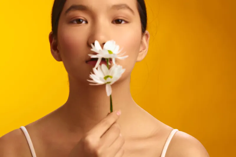 asian woman holds a white flower near her lips against a warm yellow backdrop, serene expression and soft lighting create a calm beauty portrait with natural charm.