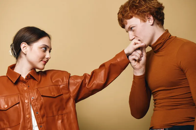Man and woman standing close indoors with affection, man kissing woman's hand, casual fashion, warm colors, romantic gesture on beige background.