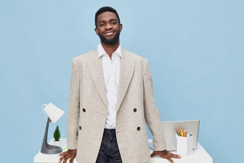 Confident smiling man standing at desk in modern office setting, wearing beige blazer and white shirt, with clean workspace including lamp, plant, and stationery, blue background, professional busines