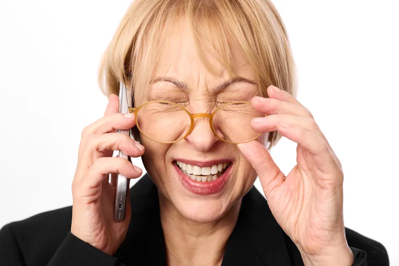 Elderly woman with glasses smiling in a close portrait, hands adjusting the eyeglass frame, phone to her ear, bright white background, lighting creates a warm mood