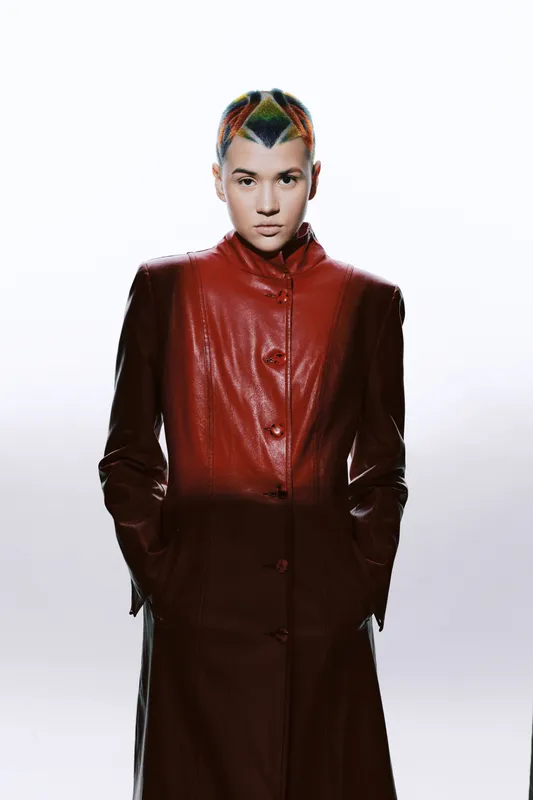 woman with colorful short hair wearing a long brown leather coat standing against a bright white background, contemporary fashion, confident expression