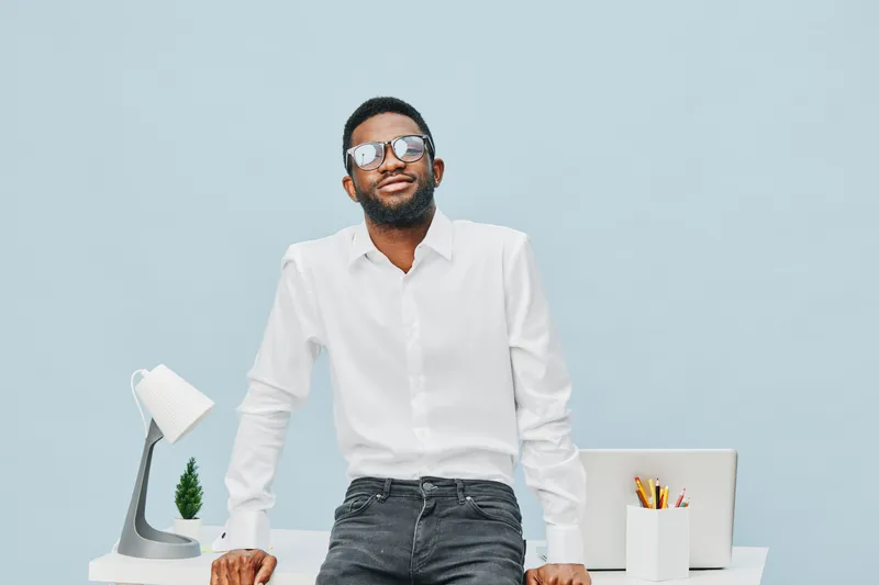 Modern confident man with beard wearing white shirt and sunglasses standing at desk in bright office space, looking at camera, relaxed and professional atmosphere, lifestyle concept