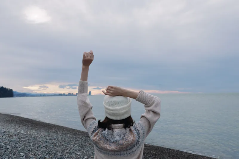 Woman with raised arms at the beach, wearing a knit sweater and beanie, facing the sea under a cloudy sky, capturing a mood of freedom and calm along the shore.