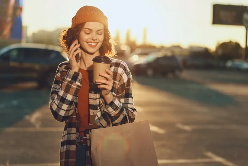 Woman with coffee and phone smiling in an urban parking after shopping, wearing a beanie and plaid coat at sunset. Authenticity, candid lifestyle, golden hour glow and mindful living.