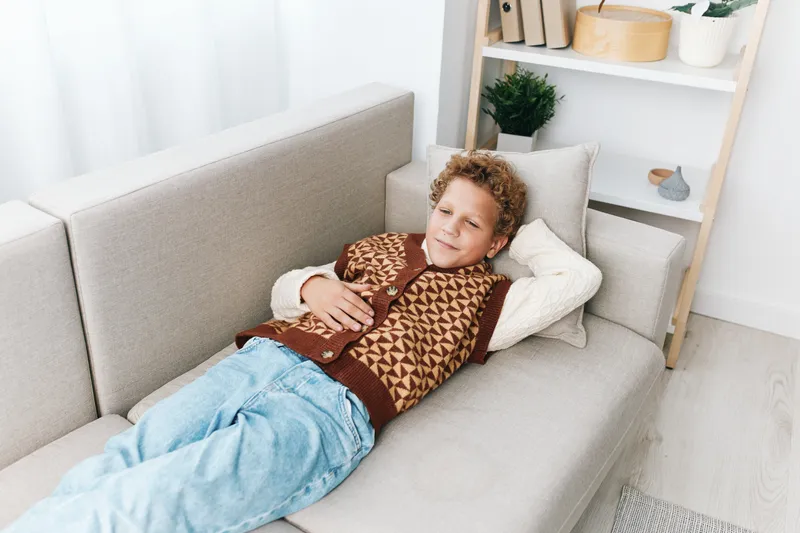 Young boy relaxes on a soft sofa wearing a knitted patterned vest, enjoying a peaceful moment at home in a bright, cozy living space.