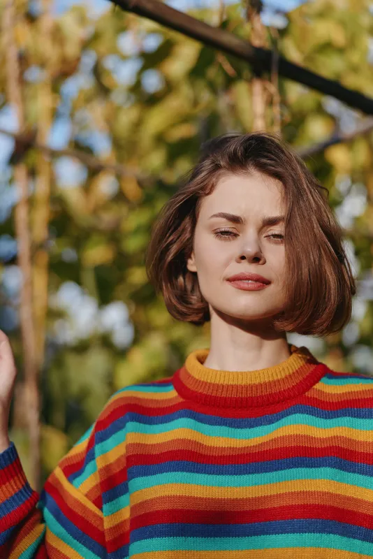 Woman in her mid 20s wearing a colorful striped sweater and bob haircut outdoors, smiling with eyes closed in sunlit nature portrait, relaxed autumn lifestyle and fashion.