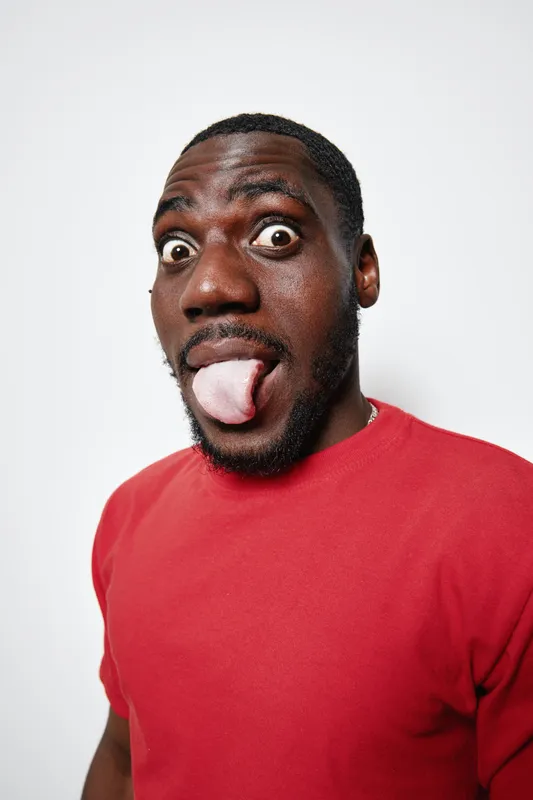 African American man with dark skin wearing red shirt making funny face with tongue out and wide eyes on white background. Portrait, expression and casual style.