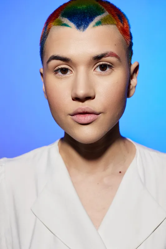 woman with colorful short hair and natural makeup wearing white collarless jacket on blue background, expressing calm and confidence with direct gaze