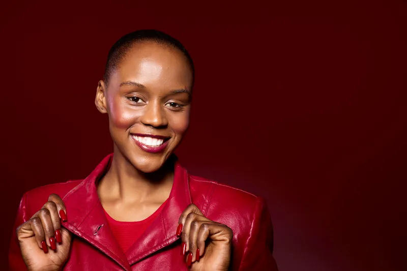 Smiling black woman with dark skin wearing a red leather jacket and matching red nails poses confidently against a deep red background. Concept for fashion, beauty, and empowerment.