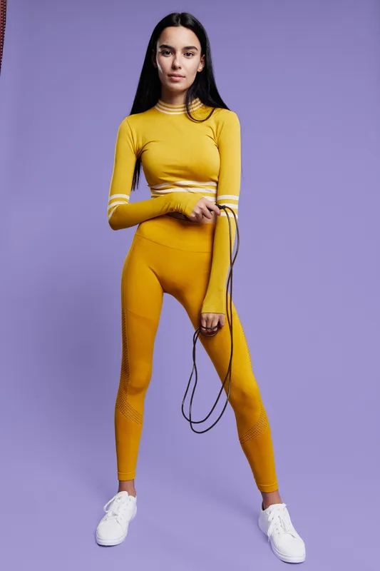 Woman sport fitness dressed in yellow outfit holding a jump rope, standing confidently against purple background, showcasing active lifestyle and modern fitness fashion.