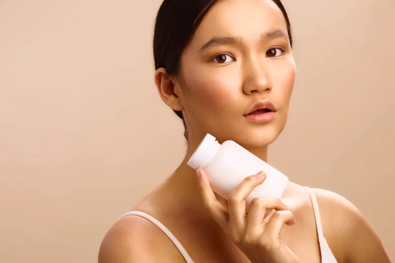 asian woman applies skincare product in a clean studio, gentle lighting highlighting smooth skin and a calm, confident mood, creating a fresh wellness vibe.